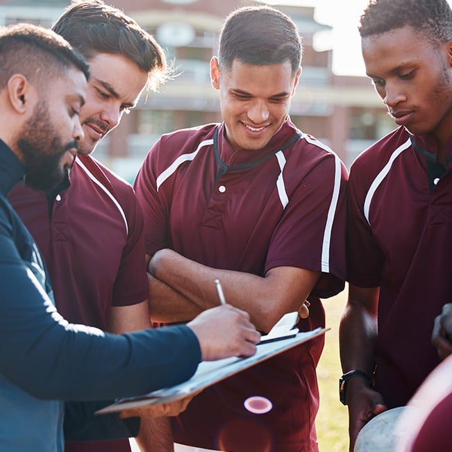 Sports team members in matching athletic uniforms gathered outdoors around a clipboard while reviewing notes and discussing information during a practice or training session.