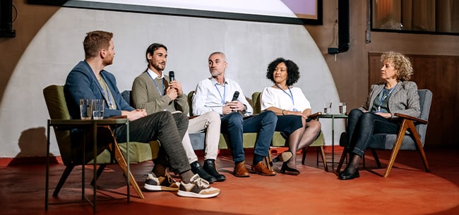 Five panelists seated on a stage during a conference discussion, holding microphones and speaking from armchairs arranged in a semicircle, with a presentation screen visible above them in an indoor event venue.
