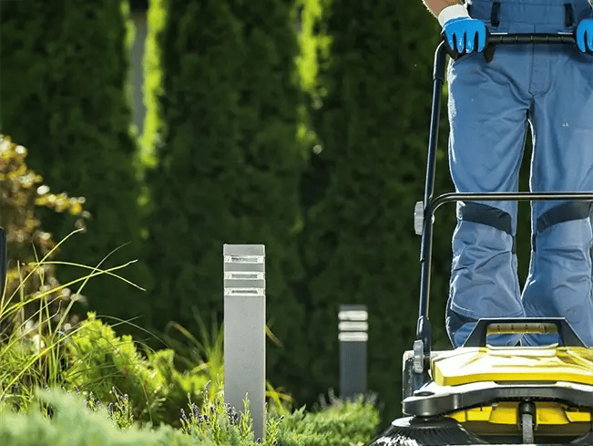Person wearing blue gloves and pants operates a yellow lawnmower in a garden with green shrubs, grass, tall trees, and two modern garden lights in the background.