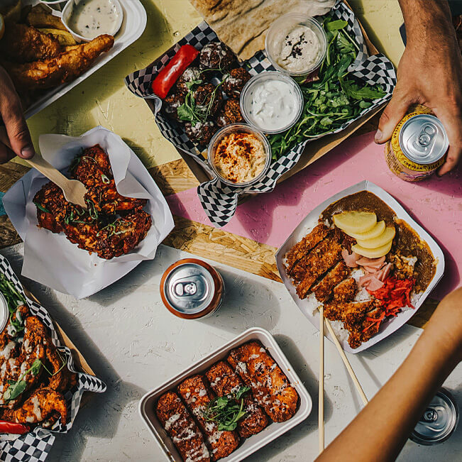 Top view of assorted takeaway dishes including grilled meats flatbread dips leafy greens and fried foods arranged on a colorful table, with beverage cans and hands reaching toward the shared meal.