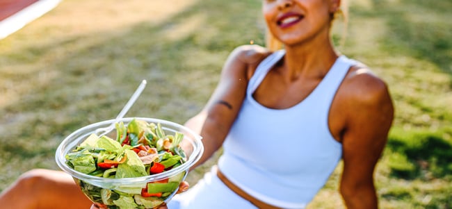 Person seated outdoors on grass holding a clear bowl of fresh salad with leafy greens tomatoes and sliced vegetables, wearing athletic clothing in a bright park setting.