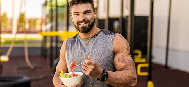 Portrait of a person in athletic wear holding a bowl of salad and a fork in an outdoor fitness setting, with exercise equipment and a modern building visible in the background.