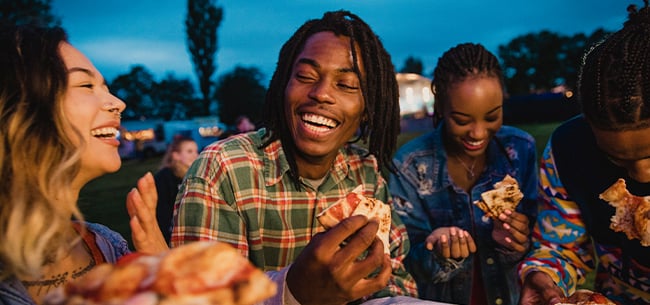 Group of people gathered outdoors in the evening, sharing pizza and food while standing together in a casual social setting with a grassy area and buildings in the background.
