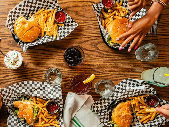 Top view of burgers and crinkle cut fries served in paper lined baskets on a wooden table, with dipping sauce soft drinks and hands holding utensils during a casual shared meal.