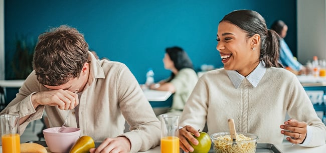 Two people seated at a dining table in a cafeteria setting, eating breakfast foods with fruit and juice, while conversing in a bright indoor space with other diners in the background.