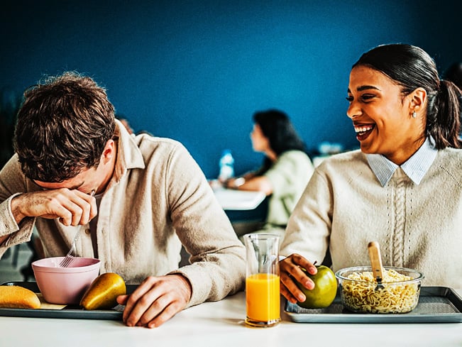 Two people seated at a dining table in a cafeteria setting, eating breakfast foods with fruit and juice, while conversing in a bright indoor space with other diners in the background.