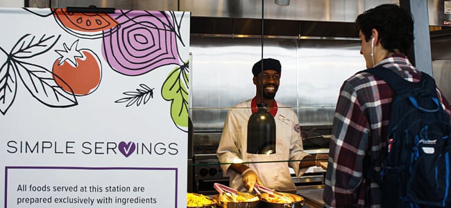 Person with a backpack ordering food at a cafeteria service counter, with a sign highlighting allergen information and simple servings, while a food service worker stands behind the serving station.