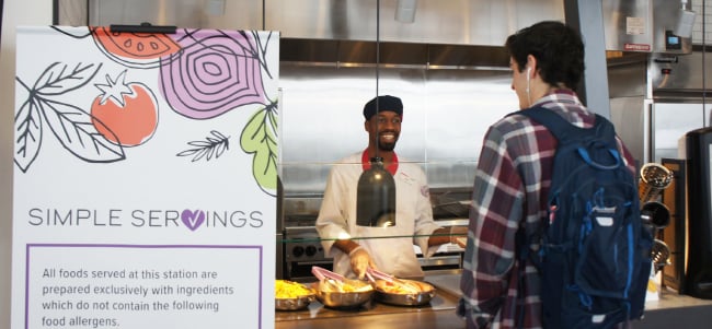 Person with a backpack ordering food at a cafeteria service counter, with a sign highlighting allergen information and simple servings, while a food service worker stands behind the serving station.