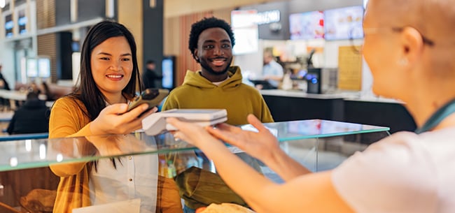 Customer paying for a boxed meal with a smartphone at a food service counter, while another customer waits nearby in a modern campus dining environment.