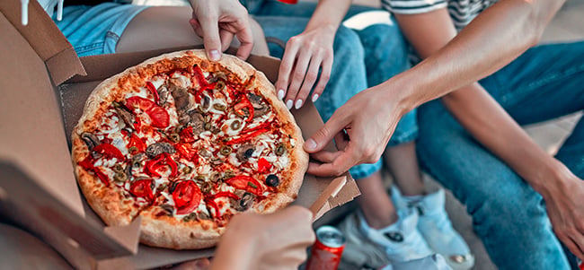 Several hands reaching into an open pizza box to share slices of pizza, with people seated nearby and a beverage can visible in a casual group dining setting.