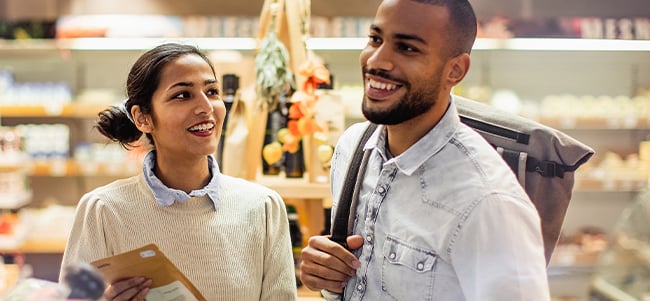 Two people standing in a retail food market, reviewing a shopping list while browsing refrigerated displays, with shelves of products and fresh food items visible in the background.