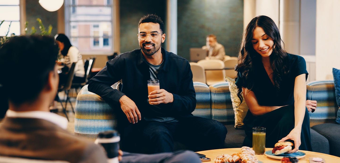 People seated on couches in a modern lounge sharing drinks and pastries on a table while talking, with additional seating and soft lighting in a contemporary indoor space.