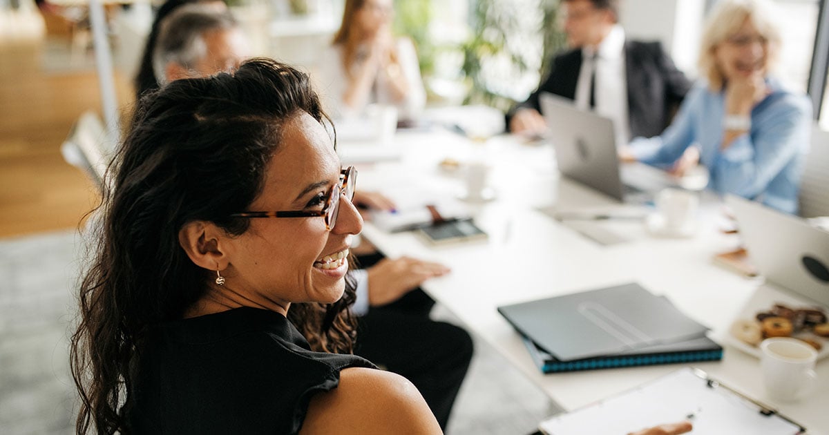Smiling woman turns toward camera during a team meeting, with colleagues and laptops blurred in background.