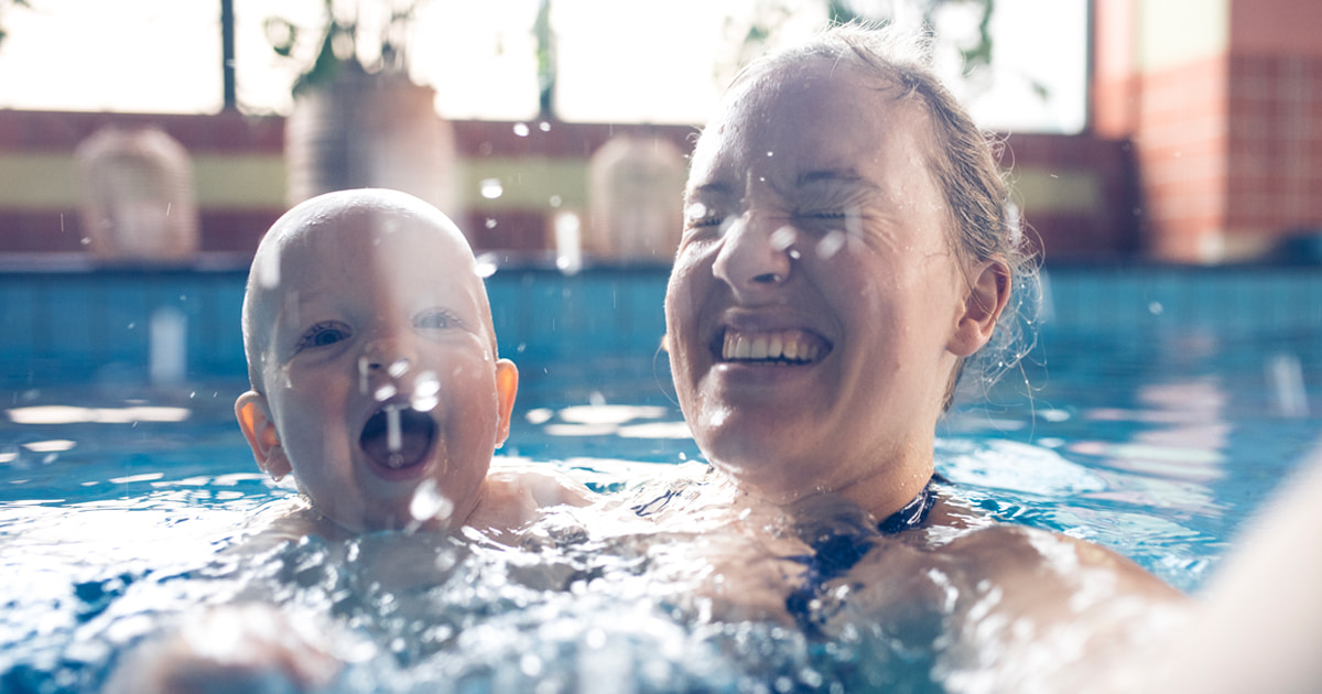 Adult holding a baby in a pool as water splashes around them. 