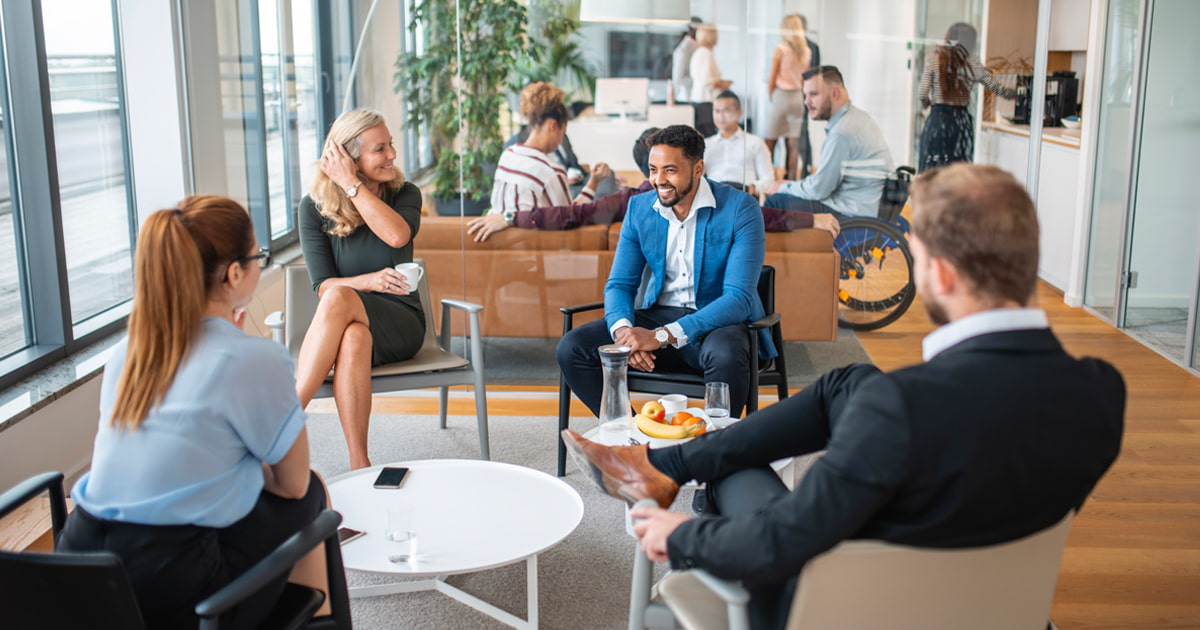 People seated in a modern office lounge having a small group discussion around a coffee table, with drinks and fruit, while others work and talk in the background.