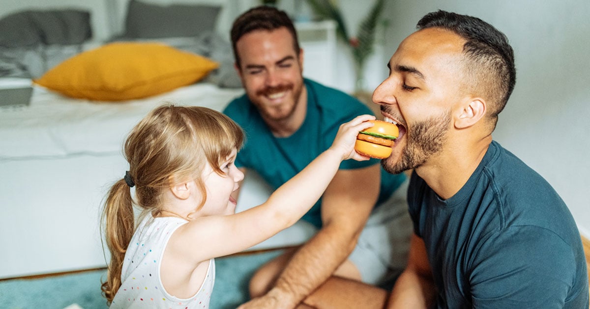 Toddler playfully offering a toy hamburger to an adult as another adult watches and smiles nearby.