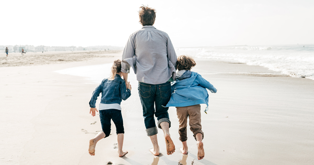 Adult and two children holding hands running barefoot along a sandy beach near the shoreline.