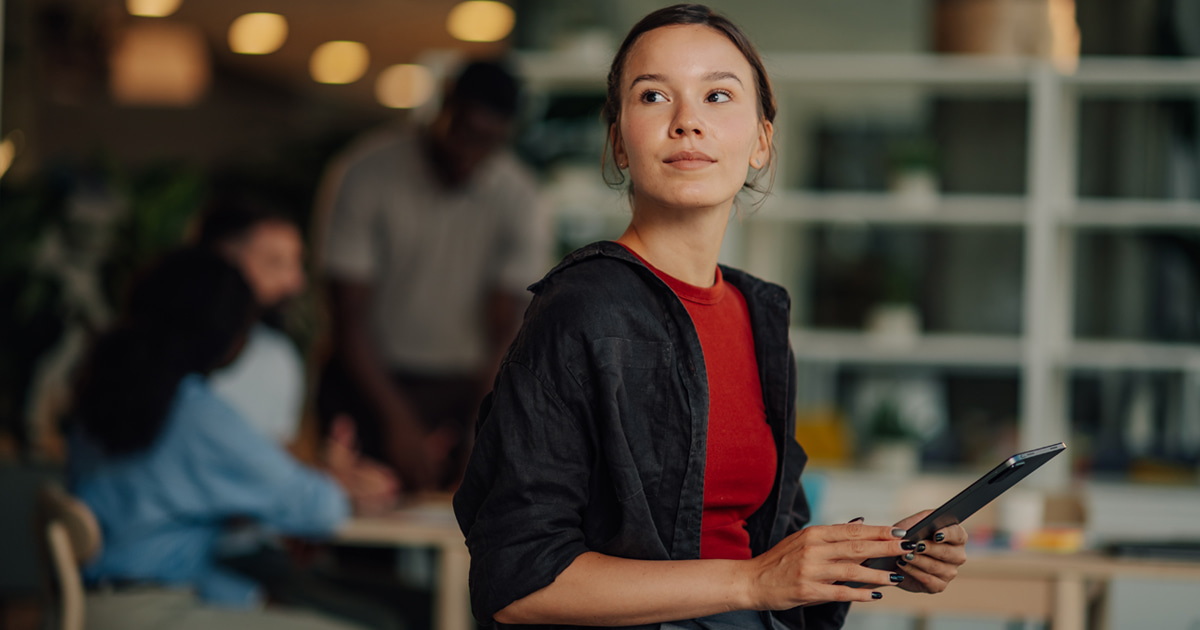 Person seated indoors holding a tablet, wearing casual clothing, with shelves and a blurred workspace in the background, suggesting a modern office or creative environment.