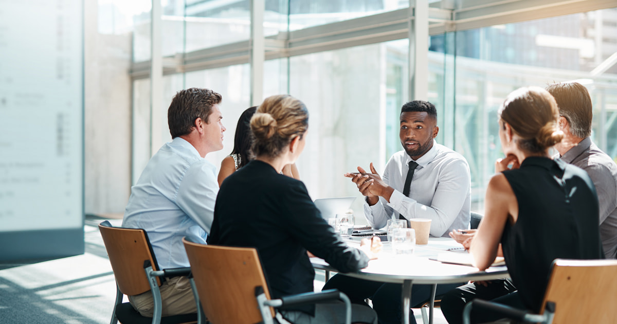 Group of colleagues sit around a table in a glass-walled office discussing during a workplace meeting.