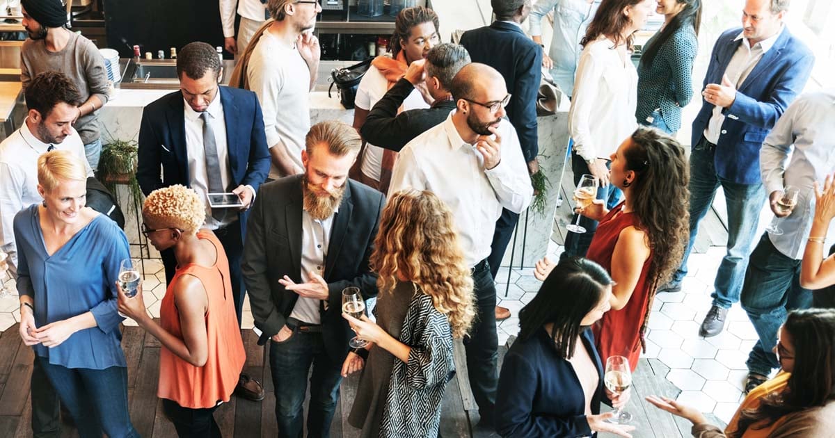 Overhead view of professionals mingling at a workplace event, standing in small groups, holding drinks, and talking in a modern indoor space with natural light.
