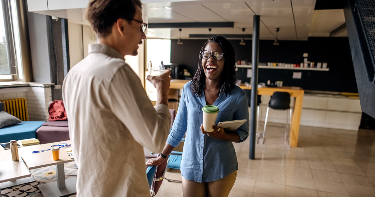 Two coworkers laughing and chatting in a modern office lounge, one holding a coffee cup and tablet.