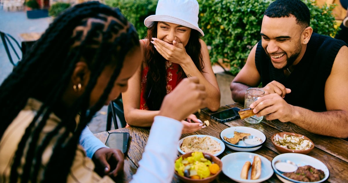 Three college friends laughing together over a shared meal outdoors.