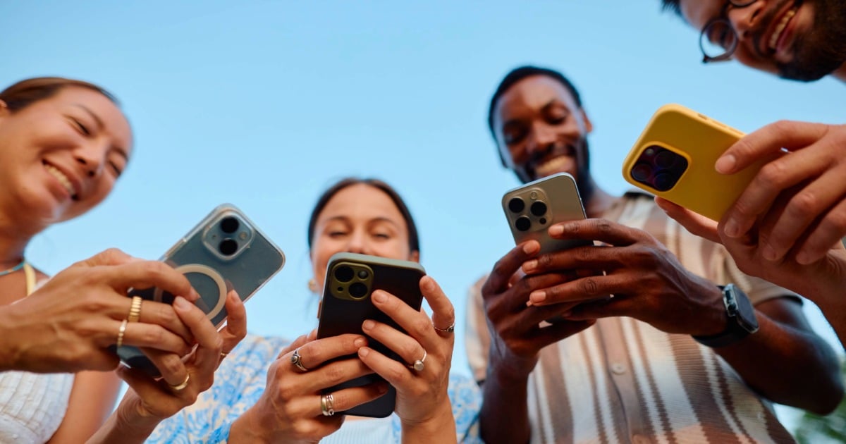 Diverse group of students gathered closely, smiling and looking at their smartphones together outdoors. 