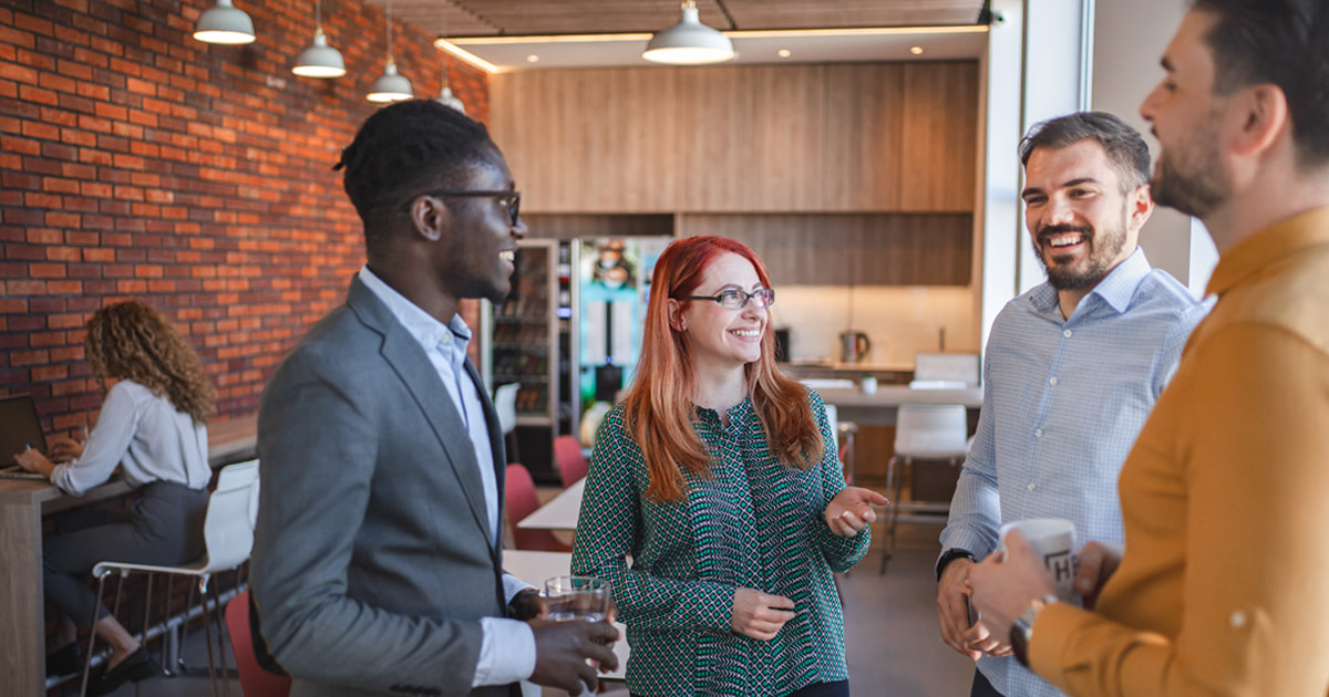 Four coworkers chat in a modern office break area, holding drinks near a kitchenette while another person works on a laptop at the counter.
