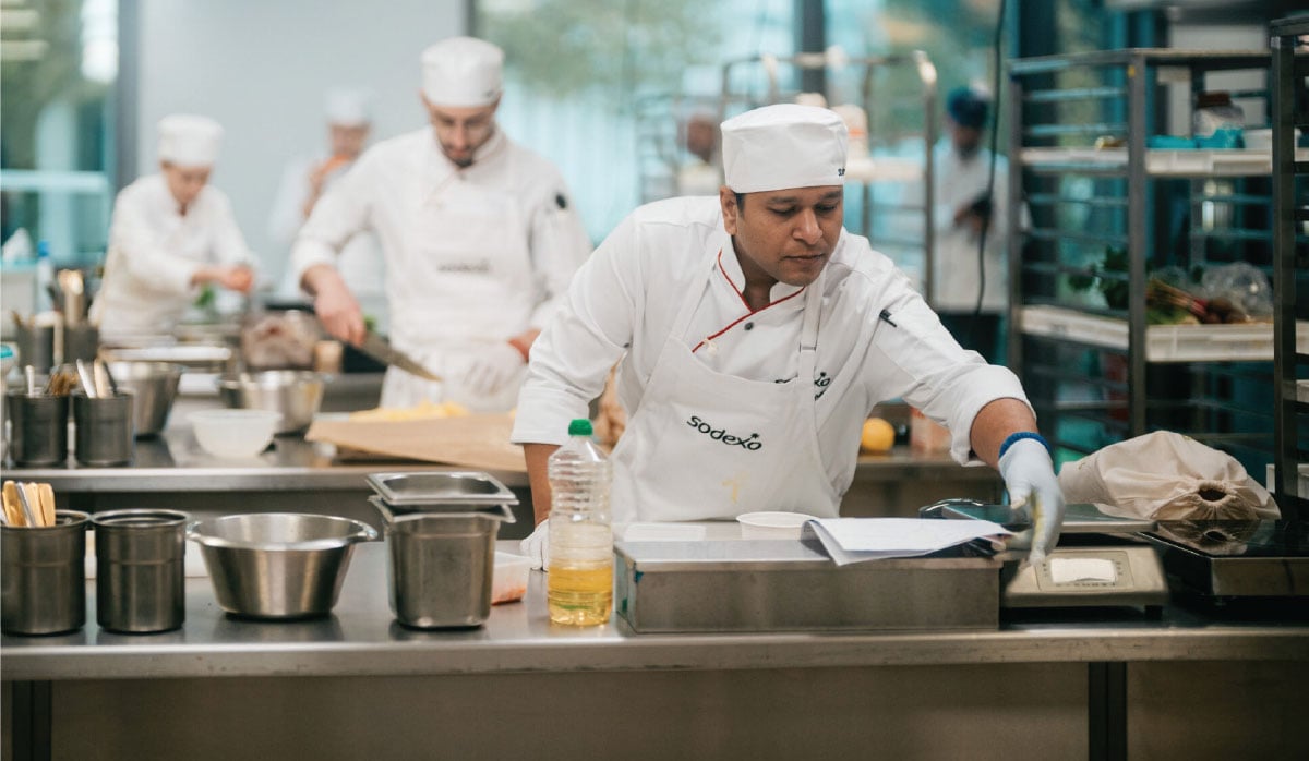 A chef in a white uniform studies a recipe sheet at a stainless‑steel workstation while others cook in the background.