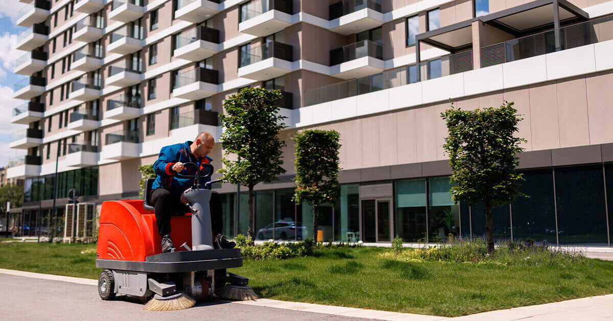 Person operating a ride-on street sweeper along a sidewalk in front of a modern apartment building.