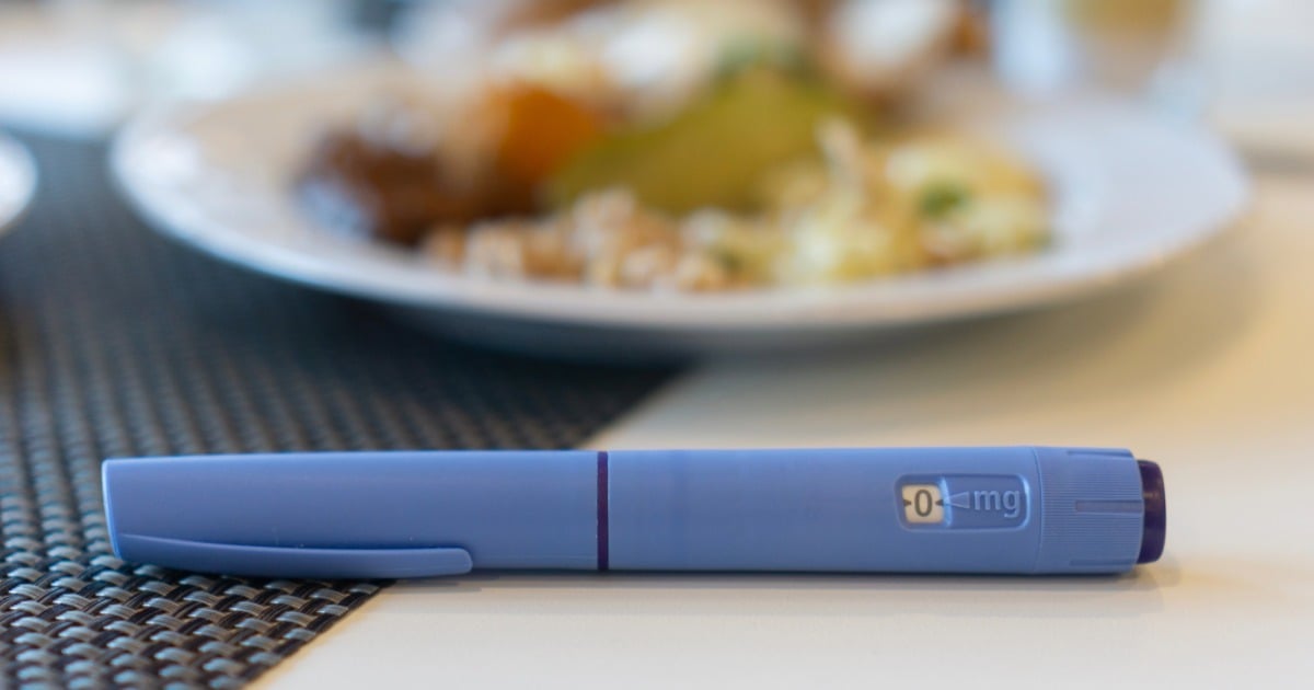 Close-up of a blue injection pen placed on a table next to a meal, with a plate of food blurred in the background, suggesting a connection between medication and dietary context.