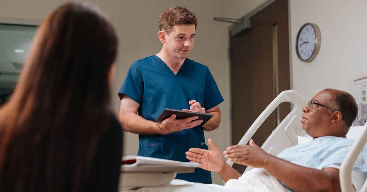 Healthcare professional in blue scrubs holding a tablet while speaking with a patient resting in a hospital bed, with another person seated nearby and medical room furnishings visible in the background.
