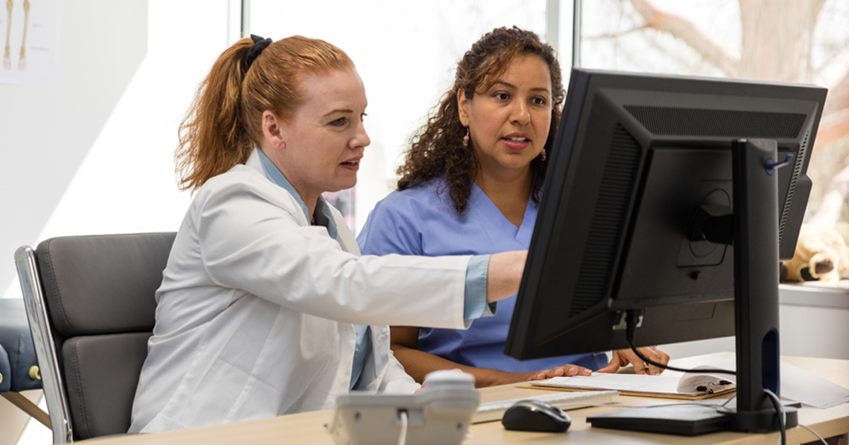 Two healthcare professionals sit at a desk in a bright medical office, looking at a desktop computer monitor.