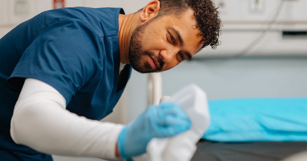 Environmental services worker cleaning and disinfecting a hospital bed with a cloth, highlighting healthcare sanitation, infection prevention, patient safety, and facility hygiene practices.