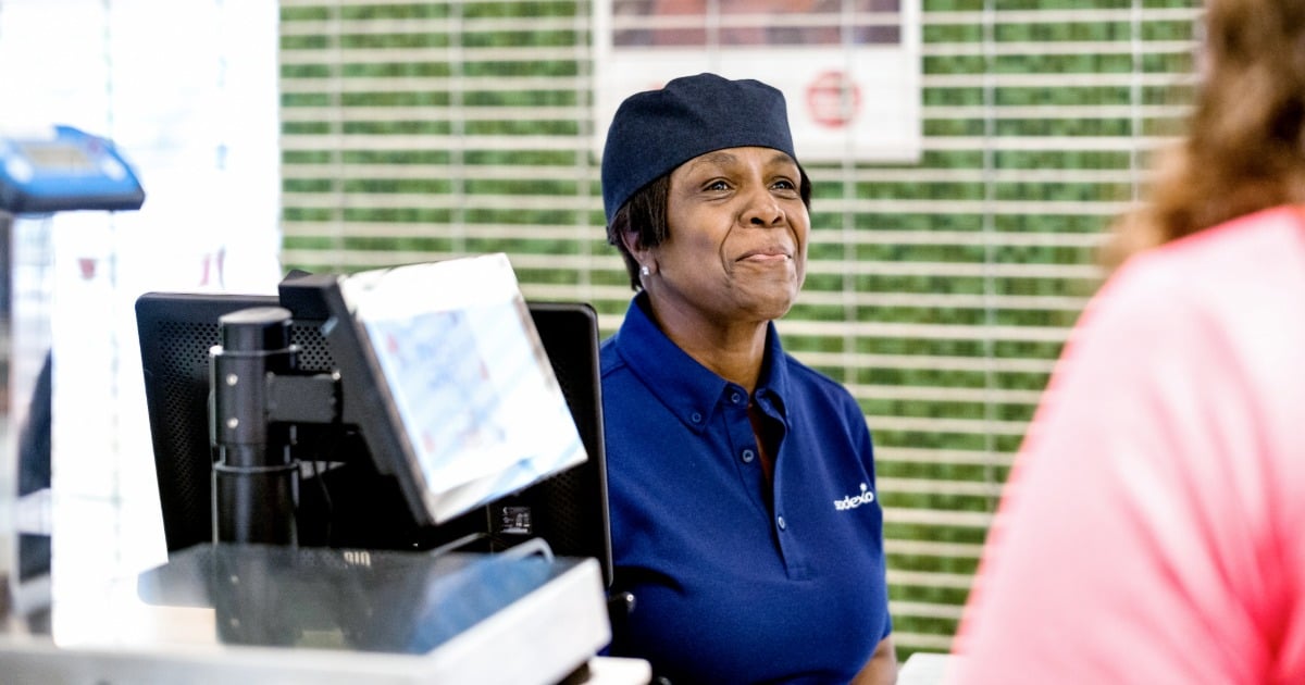 A smiling foodservice worker stands behind a register, engaging with a customer.