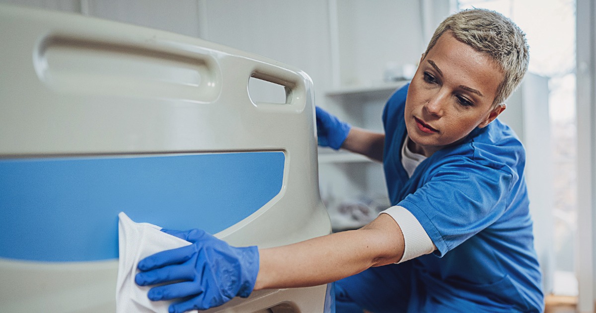 Healthcare worker in blue scrubs and gloves disinfecting a hospital bed.