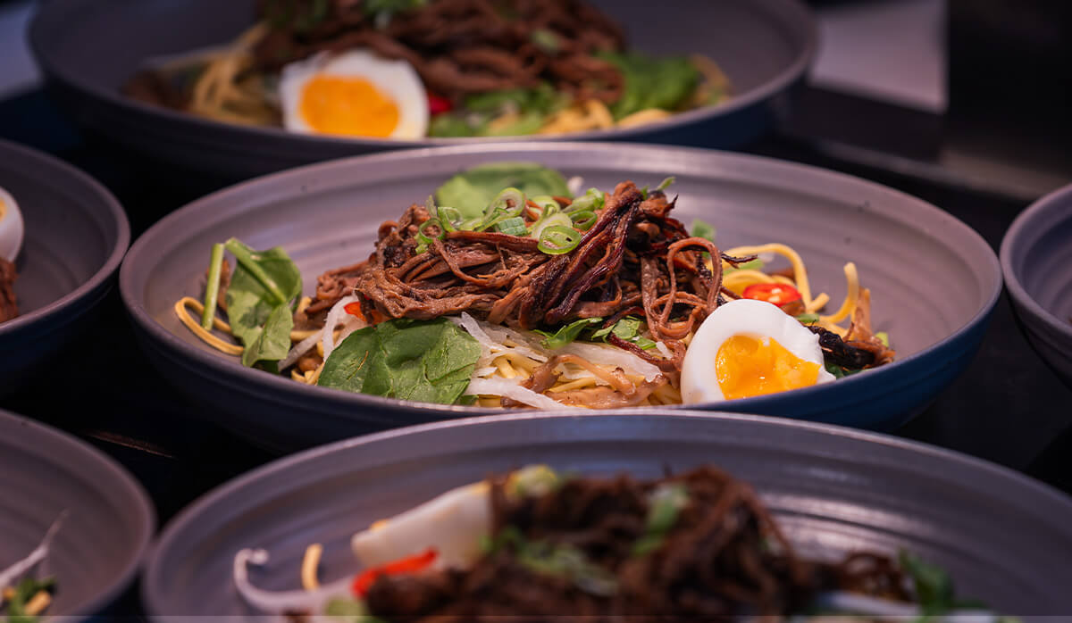 Bowl of noodles topped with shredded meat, vegetables, and a soft-boiled egg, with similar bowls in the background.