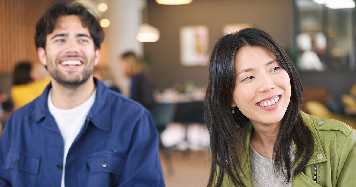 Two people seated in a café, facing slightly right, with blurred tables and warm lighting in the background.
