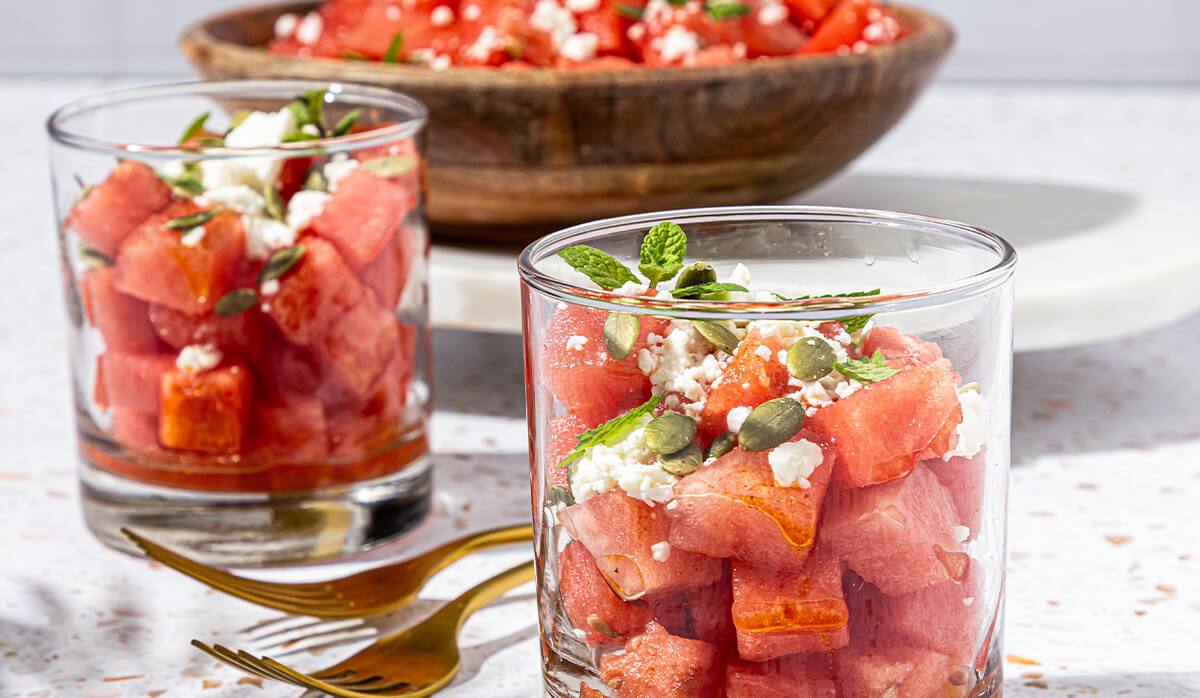 Two glass cups filled with watermelon salad topped with crumbled cheese mint leaves and pumpkin seeds, with a large serving bowl in the background on a bright tabletop.