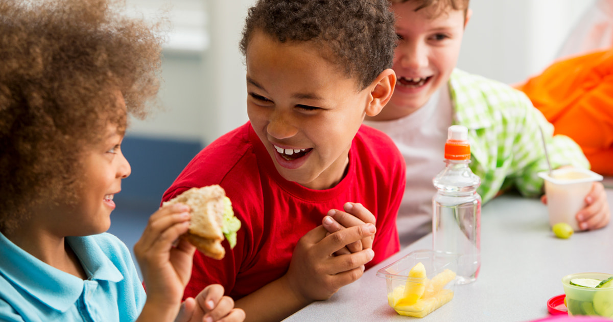 A group of three kids share a friendly moment during lunch, surrounded by healthy foods including fruit cups, yogurt, and a sandwich.