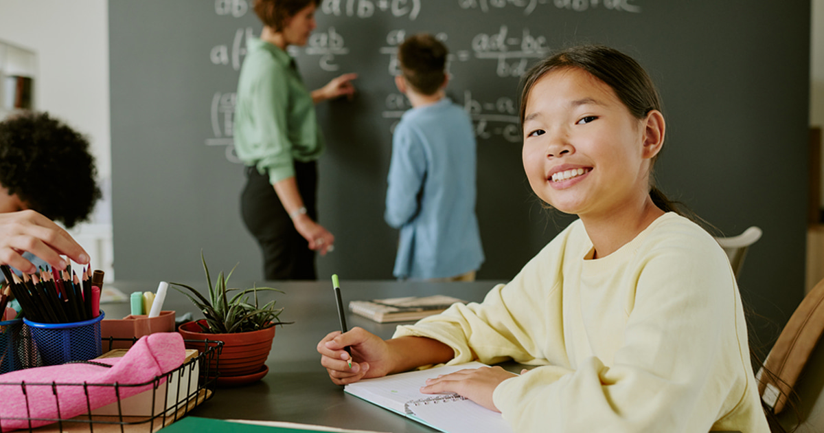 Student seated at a classroom table writing in a notebook, with pencils and school supplies nearby, while an instructor and another student work at a chalkboard in the background.