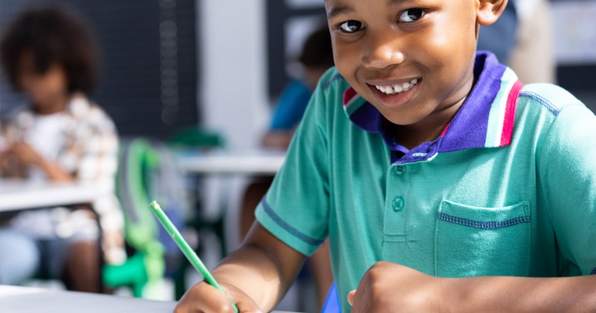 Child sitting at a classroom desk writing with a green pencil, with other students and desks visible in the softly focused background of a bright learning environment.