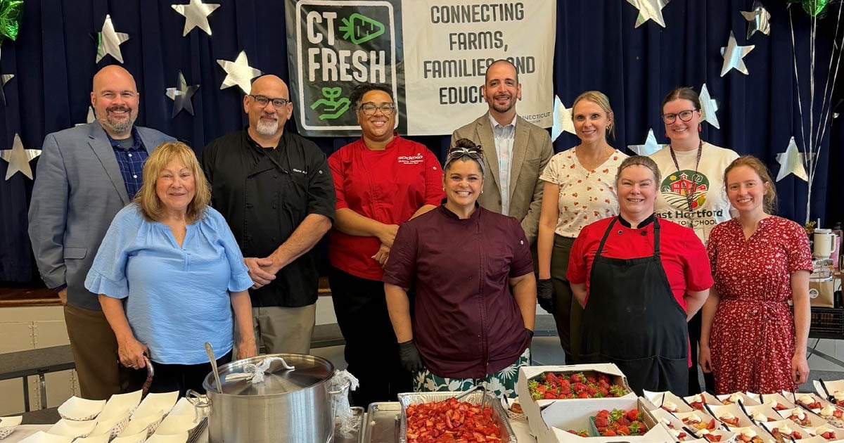 School nutrition professionals, educators, and community partners gathered around a table displaying fresh strawberries and prepared food samples at a farm-to-school event promoting local food and student wellness.