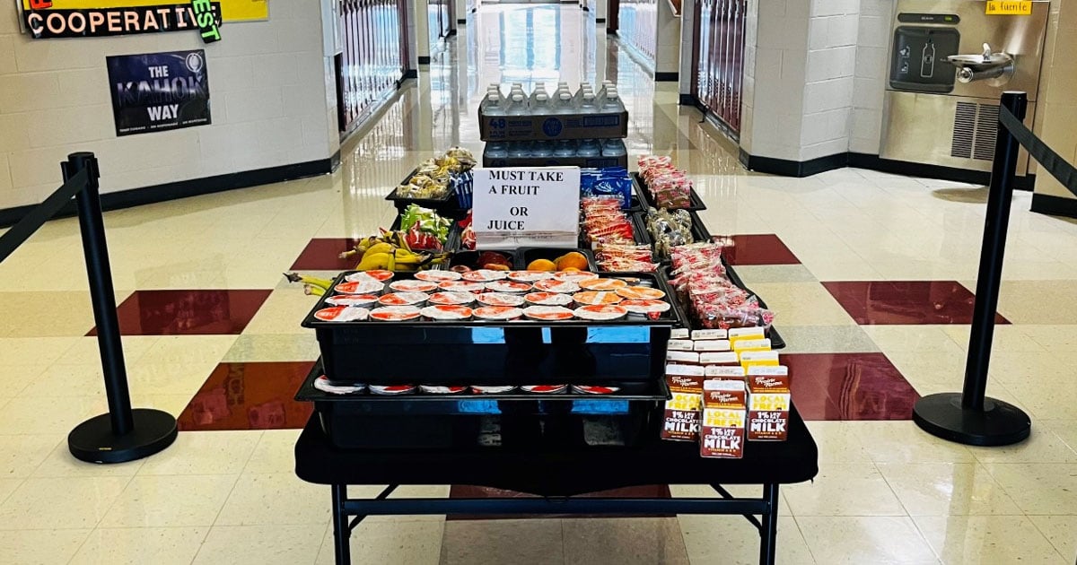 A table in a school hallway displaying fruit cups, bananas, oranges, milk cartons, water bottles, and snacks, with a sign that reads MUST TAKE A FRUIT OR JUICE.