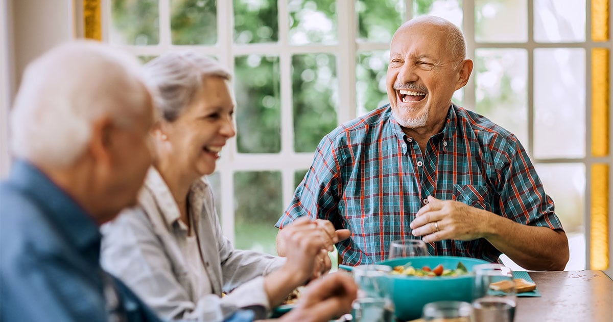 Older adults gathered around a dining table, sharing a meal in a bright room with large windows.