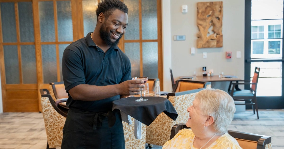 A smiling server delivers a drink to a resident in a senior living community dining room.