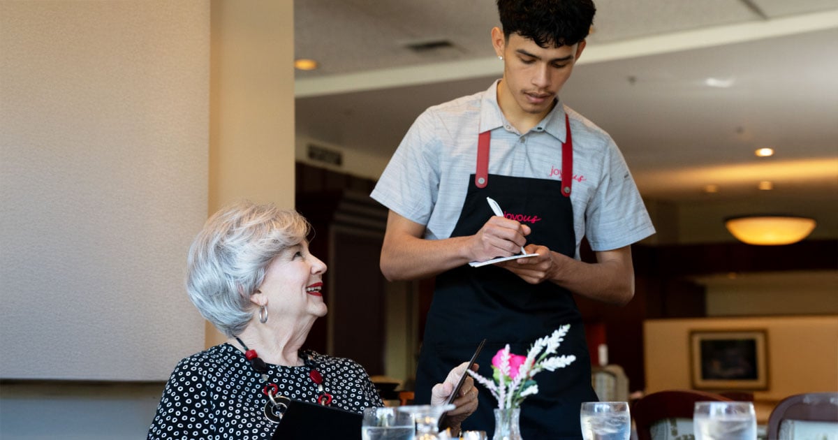 Attentive server looking down at a notepad while taking a senior resident’s order, as the resident looks up and smiles. 