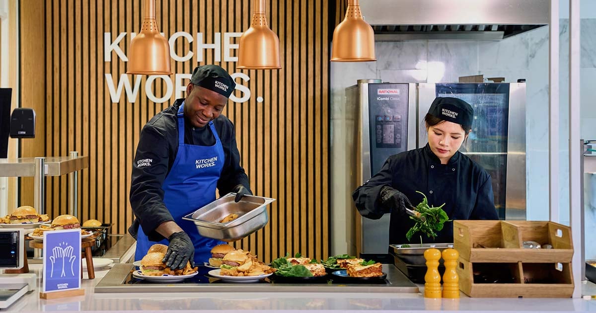Two kitchen staff preparing burgers and salads at a restaurant counter beneath Kitchen Works signage.
