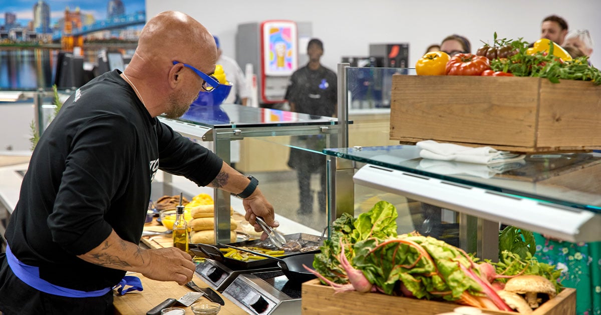 Chef preparing food at a cafeteria serving station surrounded by fresh vegetables, breads, and ingredients, highlighting made-to-order dining, culinary expertise, and nutritious food service options.