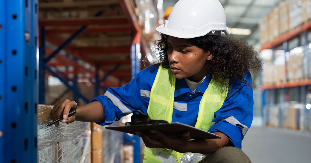 Warehouse worker in safety gear inspecting packaged inventory with a clipboard in a distribution center, highlighting facility management, inventory control, logistics, and operational efficiency.